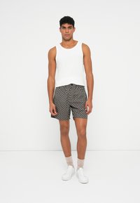 Top bianco senza maniche, pantaloni corti scuri a fantasia e sneakers bianche. I pantaloni corti presentano un motivo floreale ripetuto di piccole dimensioni. Calzini rosa chiaro visibili.