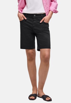 Person iført sorte knælange shorts, hvid skjorte, lyserød stribet langærmet skjorte og sorte slidssandaler, hænder i lommerne, stående.