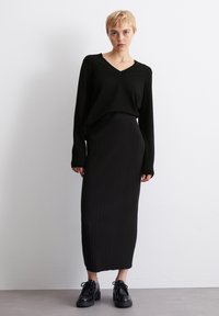 Svart strikket genser med V-hals, kombinert med en svart ribbestrikket midikjole. Modellen har på seg svarte snørebåndsplattform sko. Minimalistisk design.
