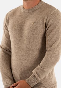 Pull en maille beige avec un col rond, des poignets côtelés et un petit logo brodé en forme de lune sur la poitrine. Texture douce et coupe classique.