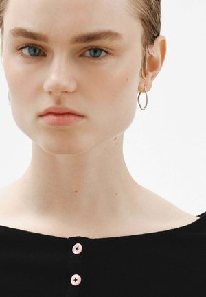 Top negro con escote redondeado, adornado con tres botones rosas. El modelo tiene el cabello corto y lleva aretes de aros plateados. La piel parece clara y suave.