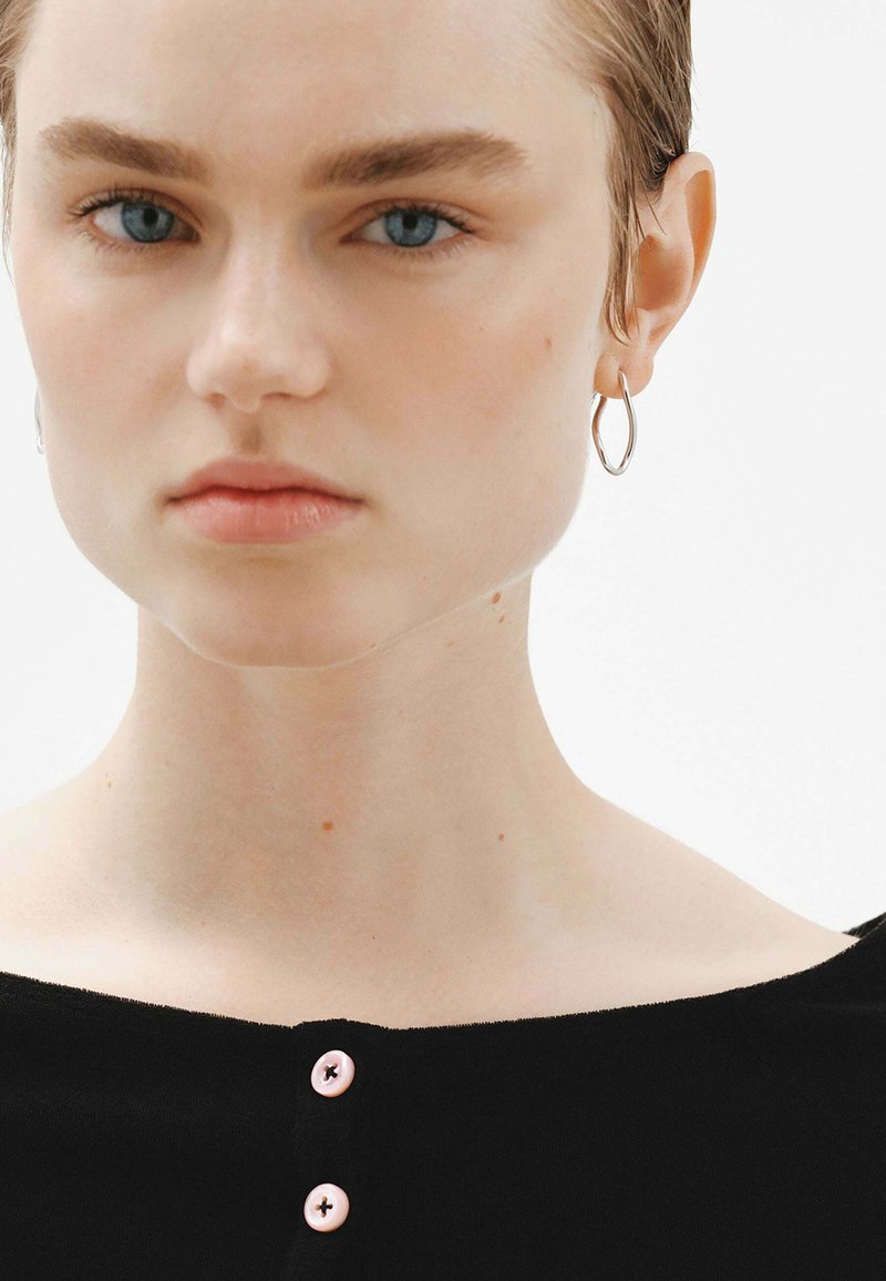Top negro con escote redondeado, adornado con tres botones rosas. El modelo tiene el cabello corto y lleva aretes de aros plateados. La piel parece clara y suave.