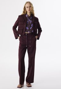 Weinrot gekürzte Jacke mit mehreren Taschen, kombiniert mit einer lila blumenmusternden Bluse und weiten weinrot gefärbten Hosen. Sandalen runden den Look ab.