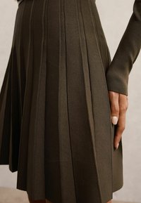 Sötét olíva zöld kötött, pliszírozott szoknya, amely kiemelkedő függőleges csíkokkal, sima textúrával és testhezálló derékkal rendelkezik.