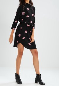 Černé šaty s růžovými květinovými vzory, tříčtvrteční rukávy, stažený pas a asymetrický lem. V kombinaci s černými kotníkovými botami.