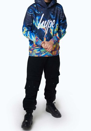 Personne portant un sweat à capuche "hype" bleu et multicolore, un pantalon cargo noir et des baskets noires, debout les mains jointes.