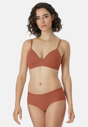 Junge Frau mit lockigen Haaren, die einen rostfarbenen BH und nahtlose Unterwäsche in passender Farbe trägt, vor einem schlichten hellen Hintergrund stehend.