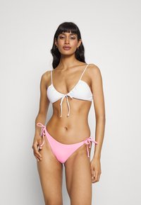Weißer strukturierter Bikini-Oberteil mit verstellbaren Bändern und einem rosa Low-Rise-Bikini-Unterteil mit seitlichen Bändern. Glatte Stoffqualität, minimalistisches Design.
