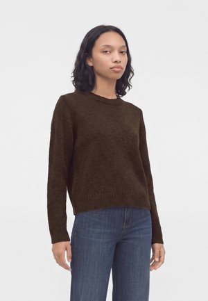 Femme aux cheveux châtains foncés mi-longs, portant un pull marron foncé et un jean bleu, debout devant un fond clair uni.