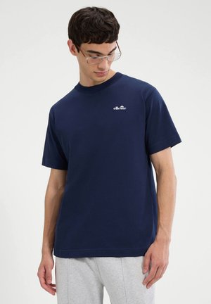 Uomo che indossa una t-shirt ellesse blu navy e pantaloni grigio chiaro, guarda verso il basso con uno sfondo neutro.