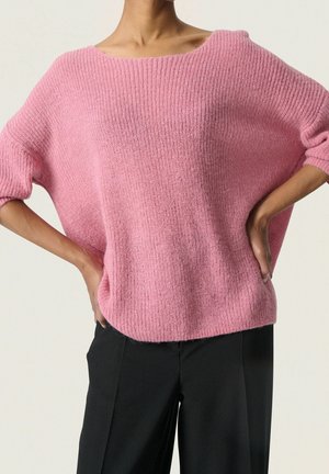 Kvinde iført løs lyserød strikket trøje med trekvartærmer, kombineret med sorte bukser, hænderne hvilende på hofterne mod en ensfarvet baggrund.