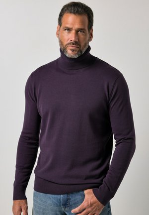 Mand med skæg iført en mørkelilla rullekrave og blå jeans mod en ensfarvet lys baggrund.