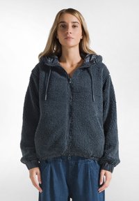 Giacca zip-up in pile grigio scuro con superficie texturizzata e cappuccio; presenta polsini e orlo elasticizzati. Design leggero e dalla vestibilità rilassata.
