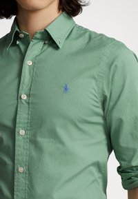 Zielona bawełniana koszula zapinana na guziki z wyraźnym kołnierzykiem. Jasnoniebieskie logo wyszywane na piersi, białe guziki oraz podwinięte rękawy.