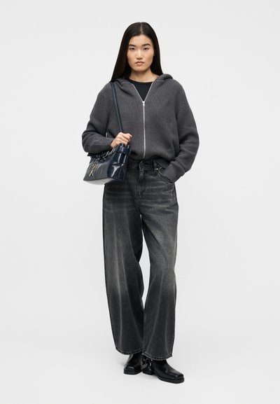 Femme portant un sweat à capuche gris en maille zippé, un jean noir délavé à jambes larges, des bottes noires, portant un sac à bandoulière noir, debout devant un fond uni.