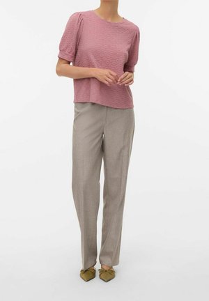 Kobieta ubrana w różowy, teksturowany krótki rękaw, beżowe spodnie o prostych nogawkach i oliwkowo zielone, spiczaste baleriny na białym tle.