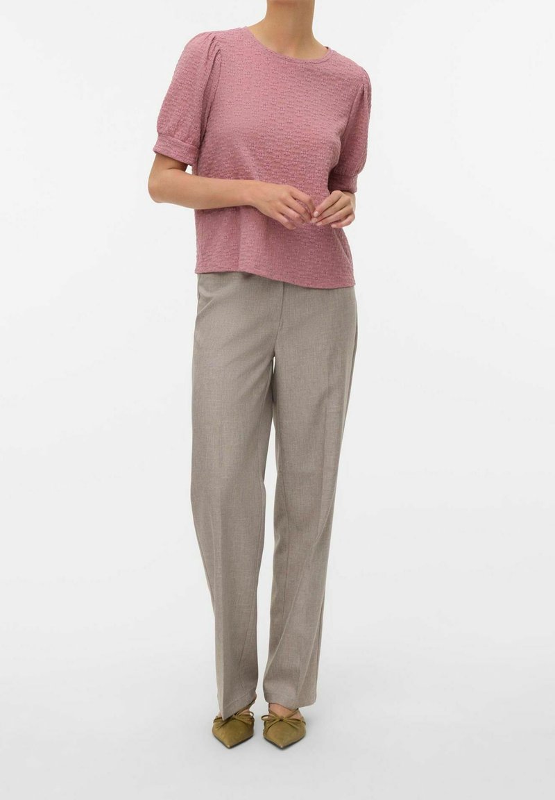 Femme portant un haut rose texturé à manches courtes, un pantalon beige droit et des ballerines pointues vert olive sur fond blanc.