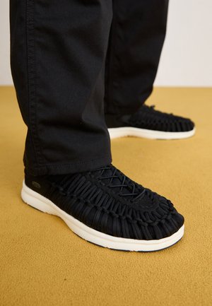 Zapatillas negras tejidas con suelas blancas en una persona que lleva pantalones negros, de pie sobre una alfombra de color mostaza.