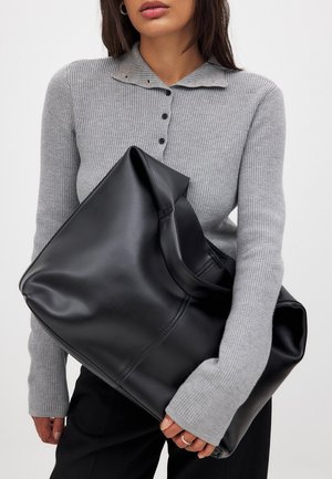 Must must nahk ülekaaluline käekott siledast tekstuurist, ümarate servade ja laia avausega, hoitud halli soonilise sviitri vastu.