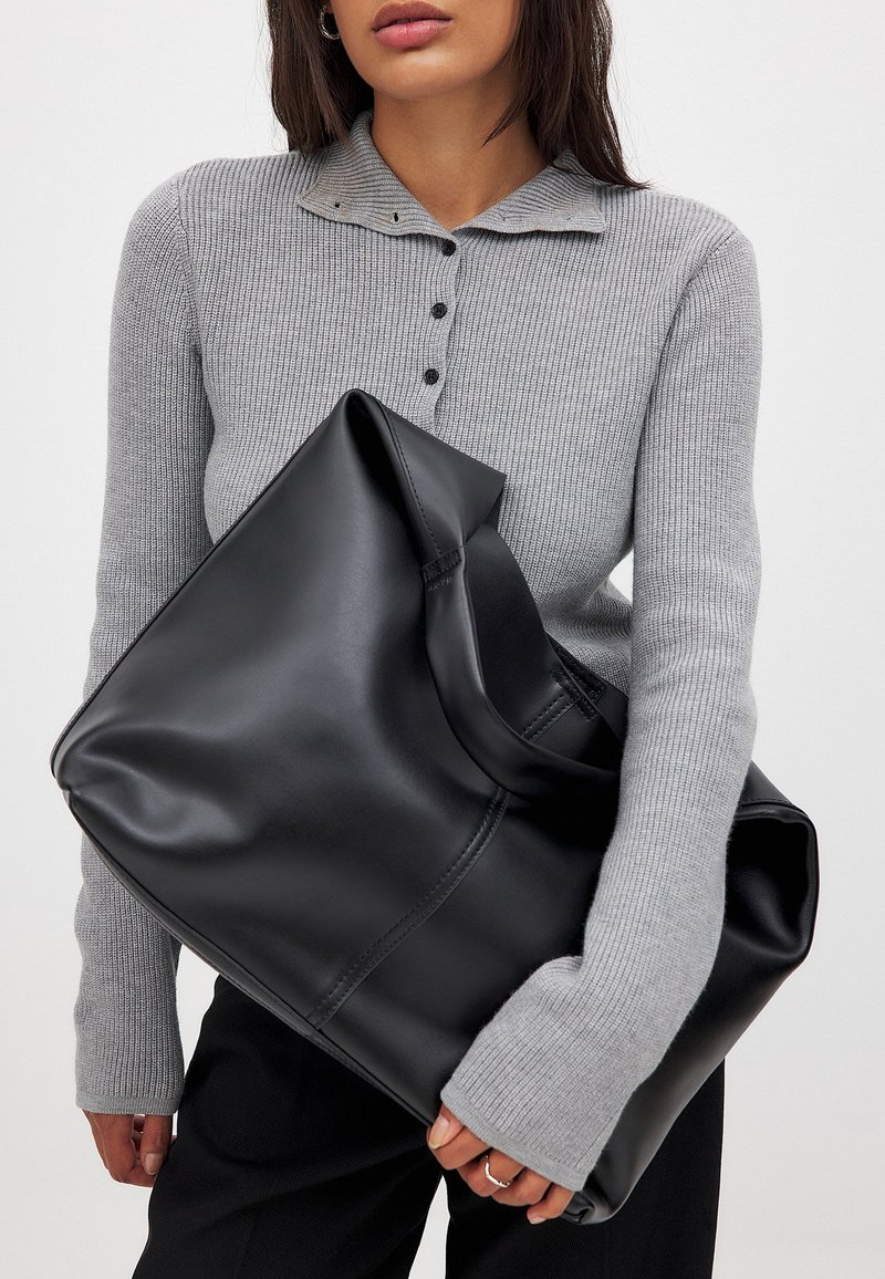 Schwarze oversized Tote-Tasche aus Leder mit glatter Textur, abgerundeten Kanten und weiter Öffnung, gehalten vor einem grauen gerippten Pullover.