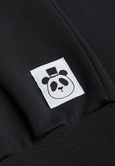 Svart stoff med en teksturert mønster; har en hvit lapp med et panda-grafikk og høyhatt, sydd på et klesplagg.