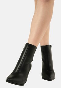 Stivali neri alla caviglia con una texture simile alla pelle liscia, punte appuntite e tacchi a cono alti. Presentano una chiusura con zip laterale per un accesso facile.