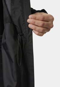 Μαύρο μπουφάν με απαλό υλικό, που διαθέτει τσέπη από δίχτυ. Ένα ρυθμιζόμενο κορδόνι είναι ορατό και κρατείται ανάμεσα στα δάχτυλα κοντά στο άνοιγμα.