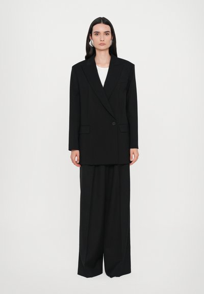 Črn oversized blazer z enim gumbom, v kombinaciji s širokimi črnimi hlačami, obema iz gladkega tekstila.