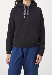 Black hooded sweatshirt with a front pocket, featuring a logo on the drawstring, ribbed hem and cuffs, paired with blue denim jeans.