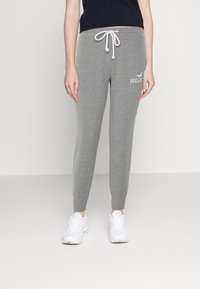 Person som bär grå dragstring-sweatpants med Hollister-logotyp och vita sneakers, stående mot en enkel ljus bakgrund.