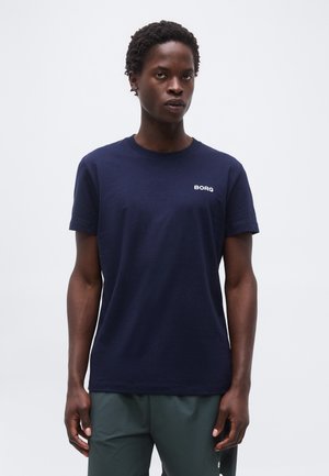 Άνδρας με κοντά σγουρά μαλλιά στέκεται αντιμετωπίζοντας μπροστά, φορώντας μπλε σκούρο t-shirt με λογότυπο BORGS και πράσινο παντελόνι, μπροστά σε ένα απλό ανοιχτό φόντο.