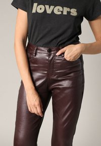 Bordeaux faux leren broek met een glanzende textuur, voorzien van voorzakken en een knoopsluiting. Het model draagt een zwart T-shirt met gouden letters.