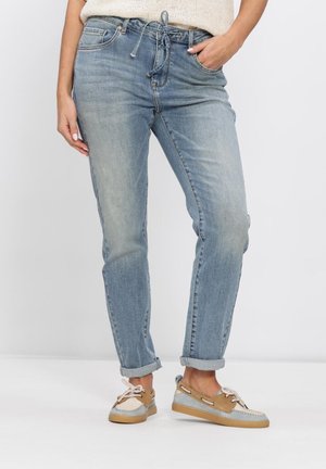 Lichtblauwe opgerolde spijkerbroek gedragen met beige en witte bootsschoenen, gecombineerd met een beige gebreide top, een hand in de zak.