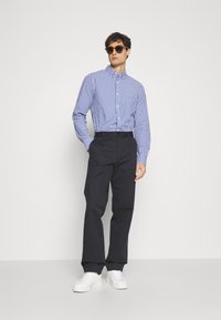 Blauw-witte geruite button-up shirt, zwarte broek, witte sneakers en ronde zonnebril. De look is casual en hedendaags.
