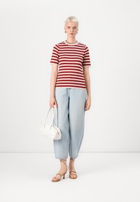 Maglietta a righe rossa e crema, jeans azzurri a taglio rilassato, borsa bianca e sandali con tacco color cuoio. Sfondo bianco semplice.