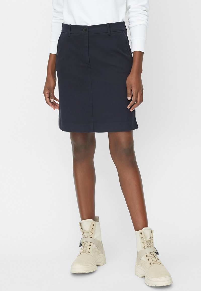 Femme portant une jupe bleu marine longueur genoux, un haut blanc à manches longues et des bottines beige à lacets, debout sur un fond blanc.