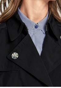 Czarny trencz z ostrym kołnierzem, ozdobiony srebrnymi elementami oraz kontrastującą, niebiesko-białą koszulą w paski pod spodem. Gładka faktura.