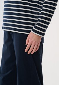 Marinblå och vit randig långärmad tröja i mjukt tyg, kombinerad med mörka byxor. Närbild av handen som vilar på överkroppen.