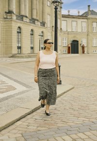 Eine Frau in einem hellrosa Tanktop und einem langen, gemusterten grauen Rock geht auf Kopfsteinpflaster in der Nähe historischer Gebäude, trägt schwarze Sonnenbrillen und Schuhe.