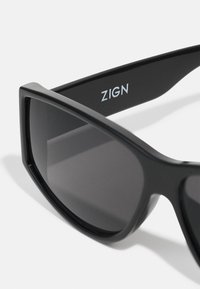 Gafas de sol negras con montura cuadrada, lentes oscuros y un acabado pulido. Presenta el logotipo "ZIGN" en plata en la patilla.