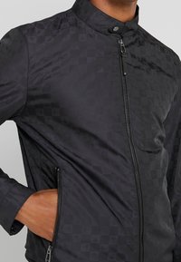 Svart jacka med ett strukturerat rutmönster, hög krage och dragkedja framtill. Har sidofickor och en slät tygfinish.