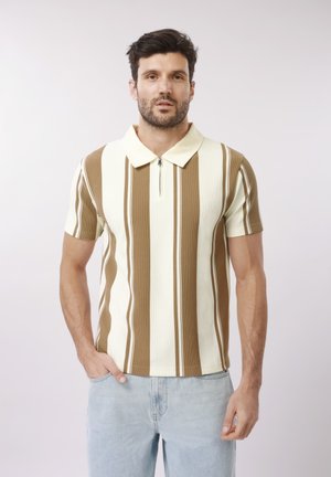 Mann mit kurzem dunklem Haar und Bart, der ein beige-weiß gestreiftes Kurzarmhemd und helle blaue Jeans trägt und mit einer Hand in der Tasche steht.