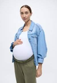Ανοιχτό μπλε τζιν μπουφάν, λευκό εφαρμοστό μπούστο, ελαστικό παντελόνι ελιάς. Γυναίκα αγκαλιάζει απαλά την έγκυο κοιλιά της, ουδέτερο φόντο.