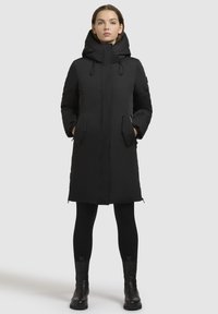 Eine Frau steht da, trägt einen schwarzen langen Daunenmantel mit Kapuze, schwarze Leggings und schwarze Knöchelstiefel vor einem einfarbigen hellgrauen Hintergrund.