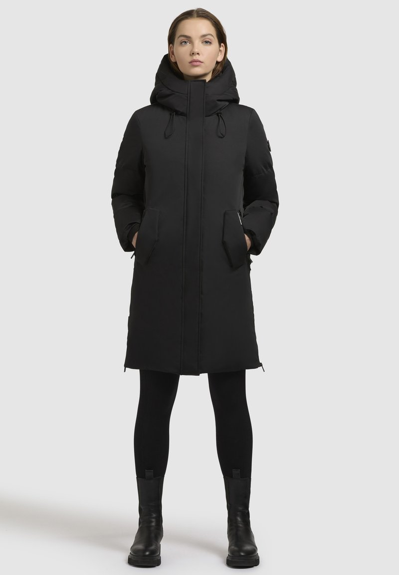 Eine Frau steht da, trägt einen schwarzen langen Daunenmantel mit Kapuze, schwarze Leggings und schwarze Knöchelstiefel vor einem einfarbigen hellgrauen Hintergrund.