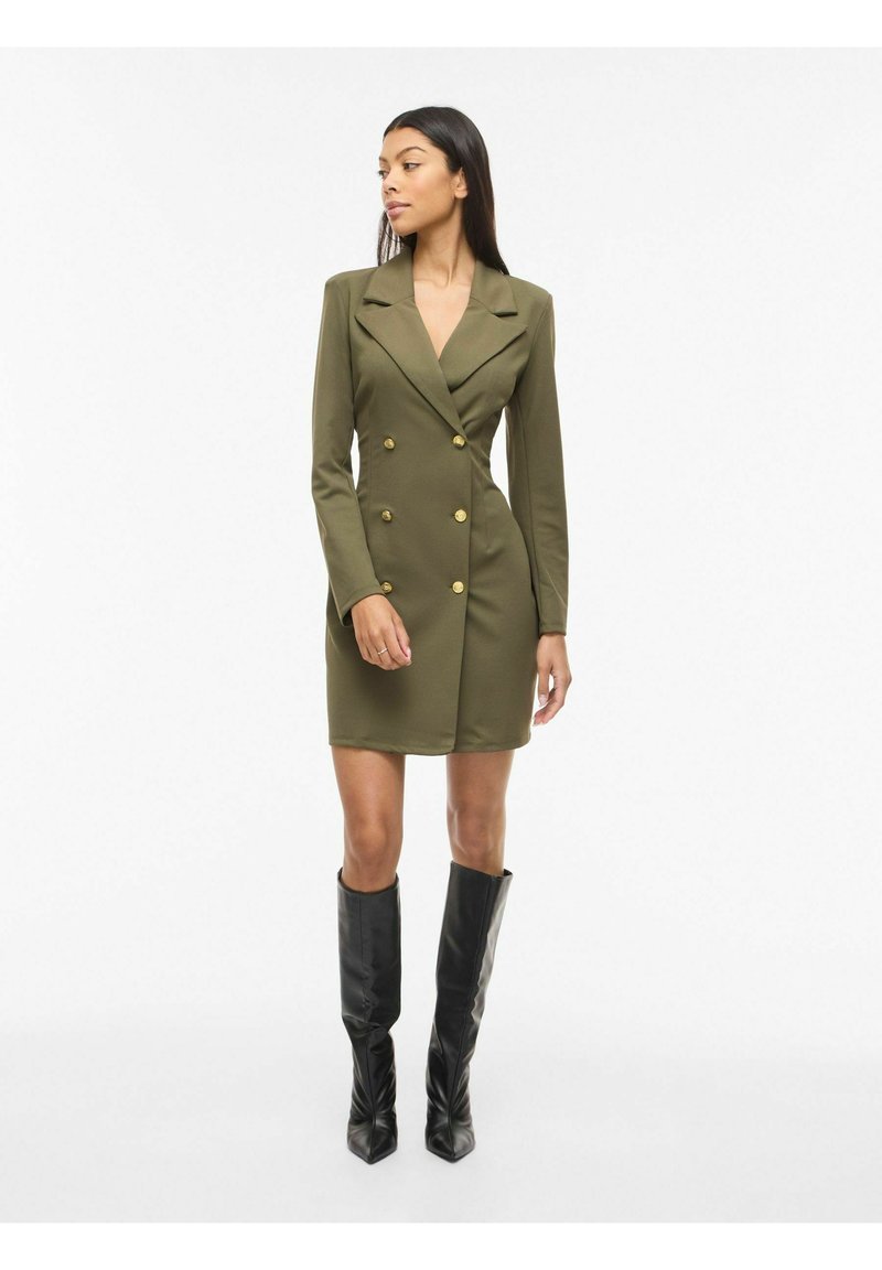 Frau trägt ein olivgrünes, doppelreihiges Blazerkleid mit goldenen Knöpfen und schwarzen kniehohen Stiefeln, steht vor weißem Hintergrund.