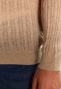 Beige kabelstickad tröja med ribbade muddar och fåll. Mjuk textur, lätt material. Mönstret har vertikal kabelstickning.