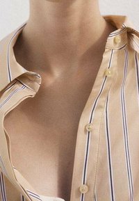 Camicia abbottonata beige con righe verticali; presenta un colletto a punta, una texture liscia e due bottoni visibili vicino al collo.