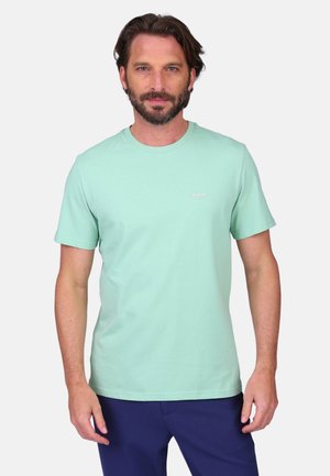 Homme aux cheveux bruns courts et barbe, portant un t-shirt à manches courtes vert menthe et un pantalon bleu marine, debout devant un fond blanc.