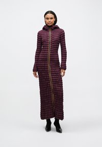 Μακριά, μωβ φουσκωτή παλτό με υφή και εμβρυαλώδη σχέδιο, κουκούλα και χρυσό φερμουάρ, που φτάνει μέχρι τους αστράγαλους. Μαύρη υπόδηση συμπληρώνει την εμφάνιση.